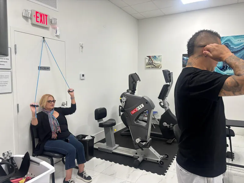 Patient doing a seated resistance band exercise beside a SciFit bike in the rehab area at All In One Specialty Health, Miami Gardens